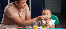 Madre alimentando con una cuchara a su pequeña hija frente a un plato con verduras y un vaso de jugo