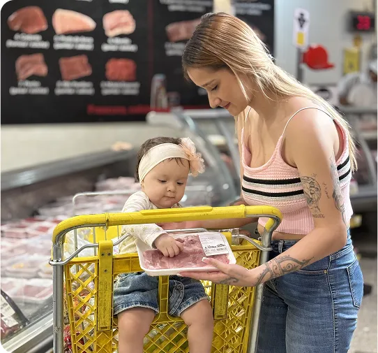 Madre mostrando a su bebé un paquete de carne mientras hacen compras en la sección de carnes de un supermercado.
