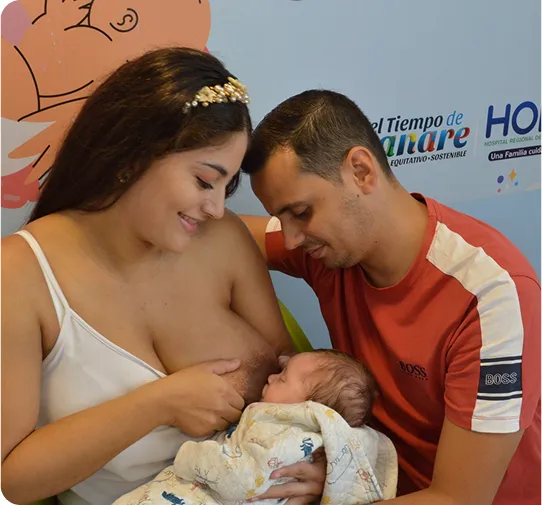 Madre amamantando a su bebé recién nacido mientras el padre observa con ternura, sentados frente a un mural promoviendo la lactancia.