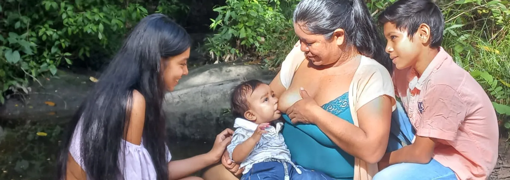 Una mujer amamantando a un bebé mientras una niña y un niño, probablemente sus hijos, la rodean con sonrisas en un entorno natural lleno de árboles.